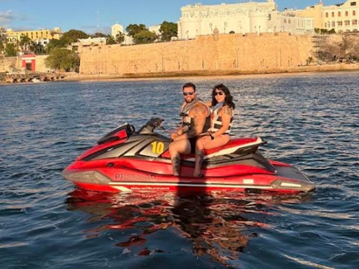 Two people on a red jet ski in a calm waterway near historic buildings.