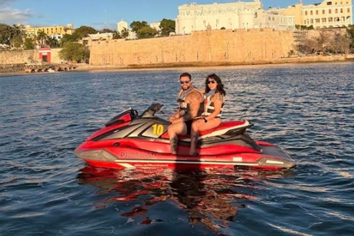 Two people on a red jet ski in a calm waterway near historic buildings.