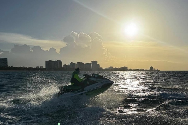 Person riding a jet ski on ocean at sunset with city skyline in the background.