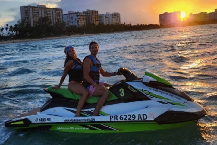 Two people on a jet ski at sunset on the ocean near a city skyline.