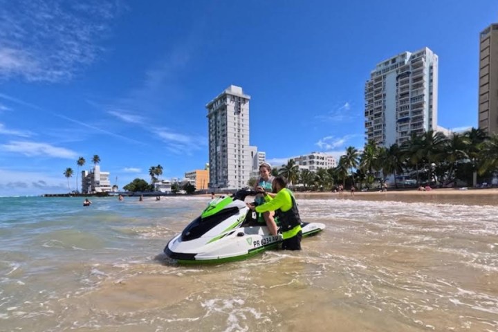 Person on a green and white jet ski near a sandy beach and tall buildings.