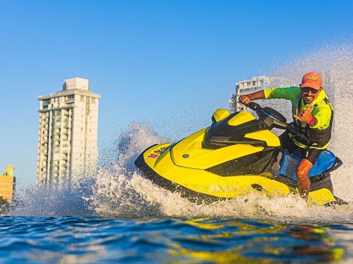 Person on a yellow jet ski riding through water near tall buildings and a clear blue sky.