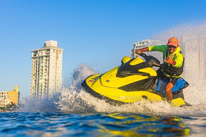 Person on a yellow jet ski riding through water near tall buildings and a clear blue sky.