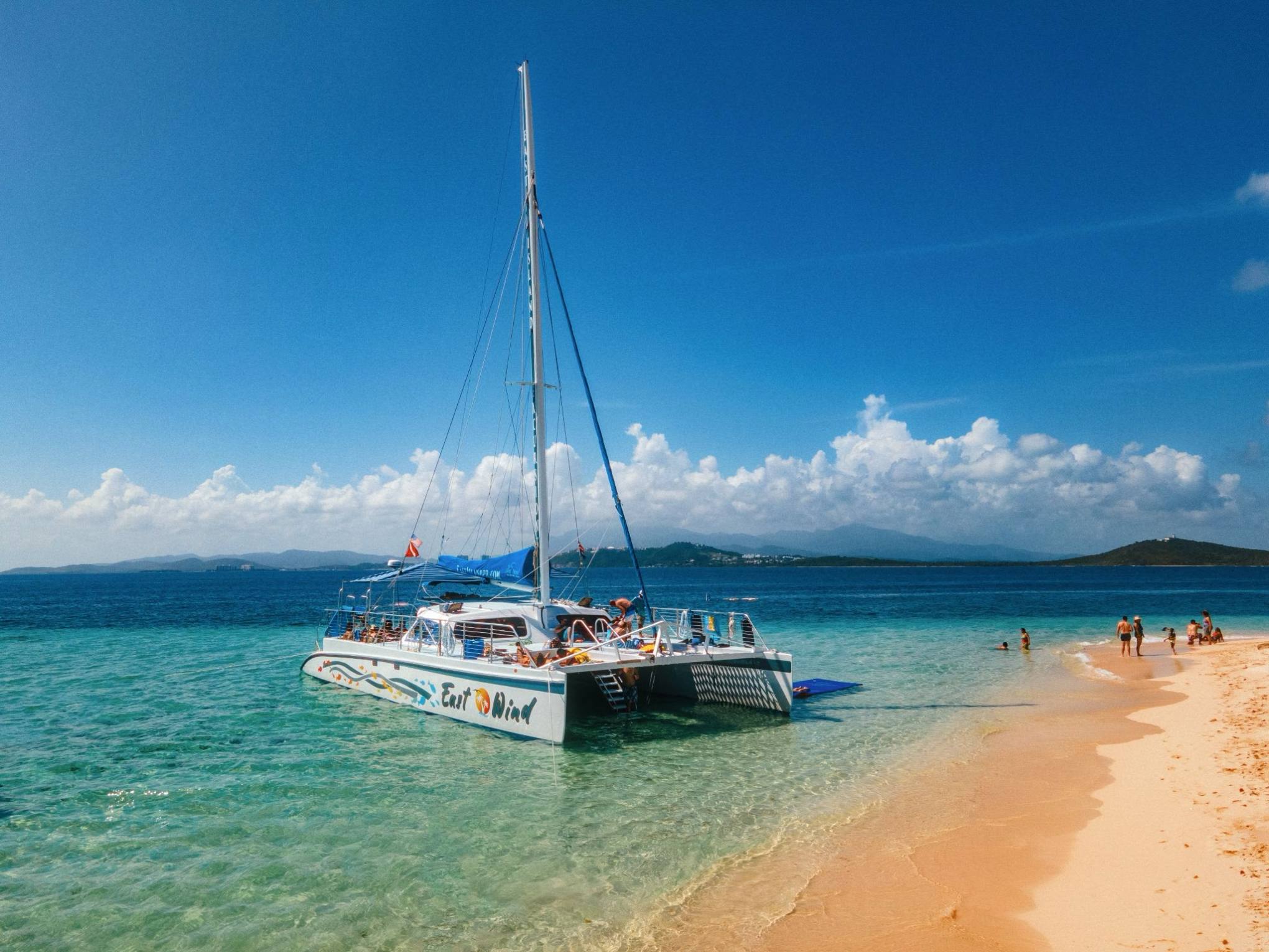 Catamaran anchored near sandy beach with people swimming.