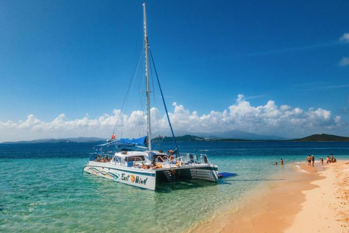 Catamaran anchored near sandy beach with people swimming.