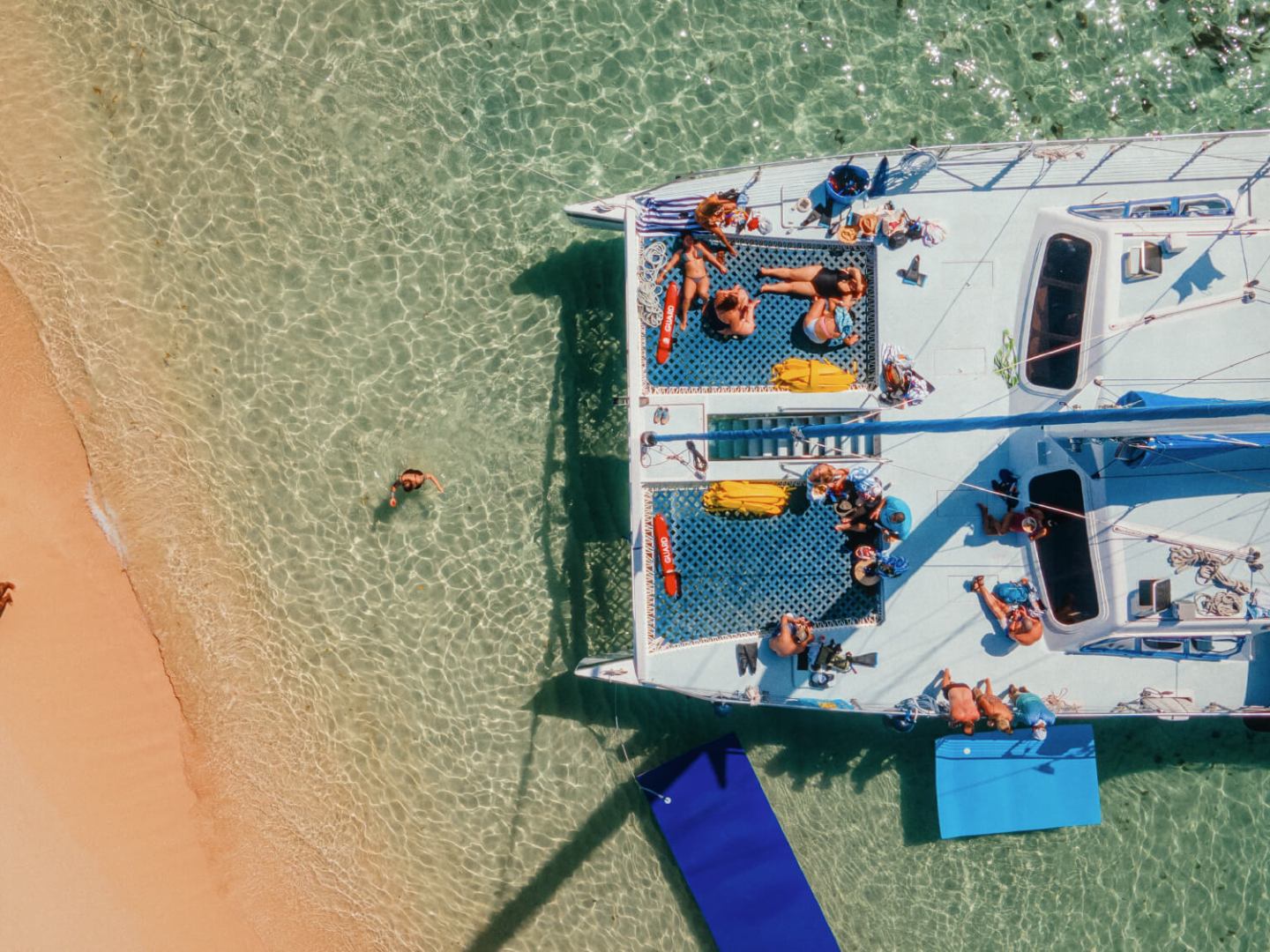 Aerial view of a catamaran with people on clear water near a sandy beach.