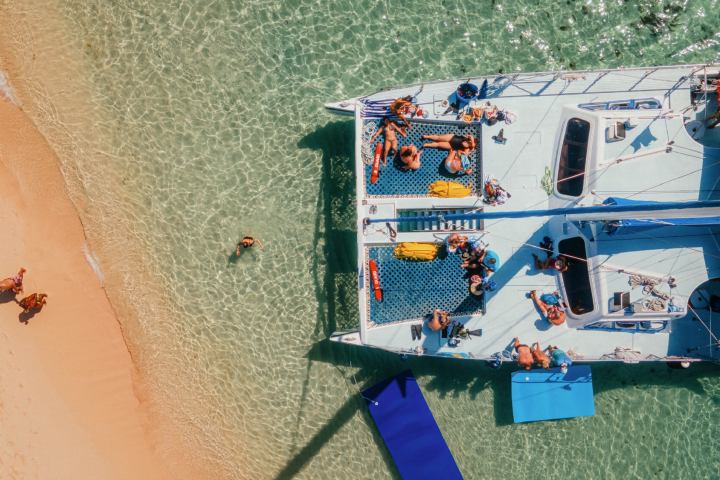 Aerial view of a catamaran with people on clear water near a sandy beach.